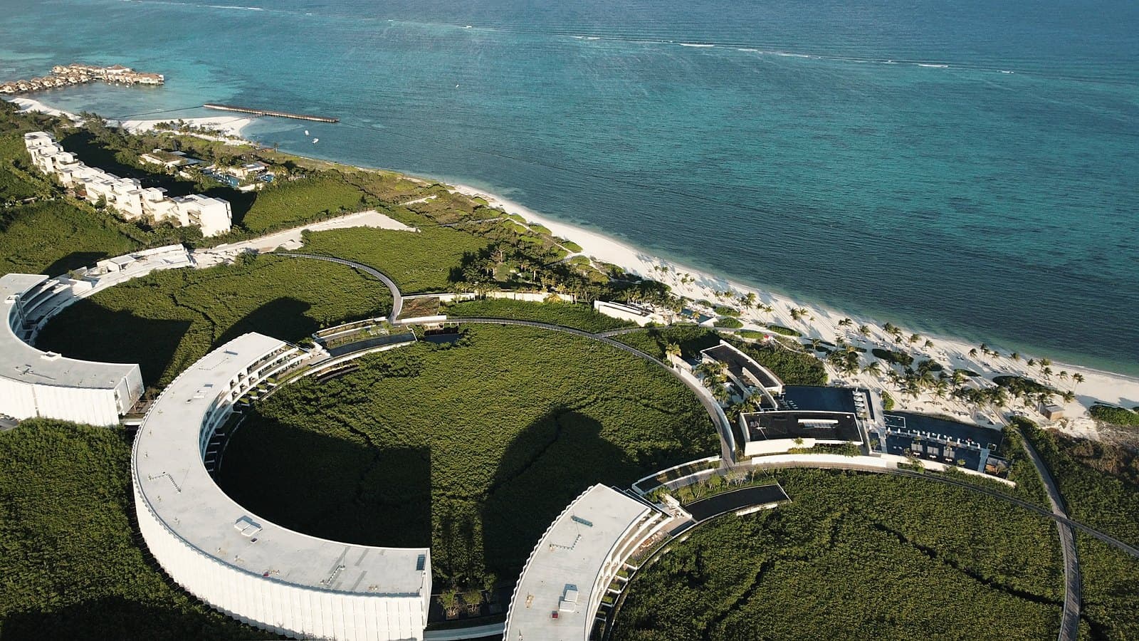 Aerial of curved green-roofed buildings running along a turquoise coastline