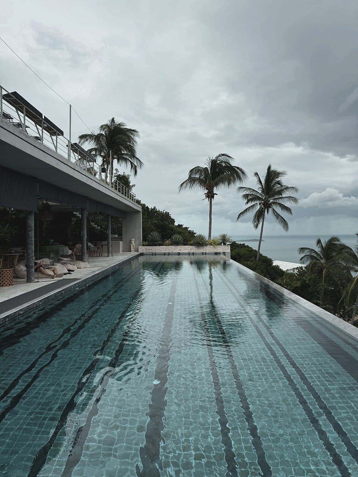 Long lap pool beside a villa terrace, palms and a grey sky at the end