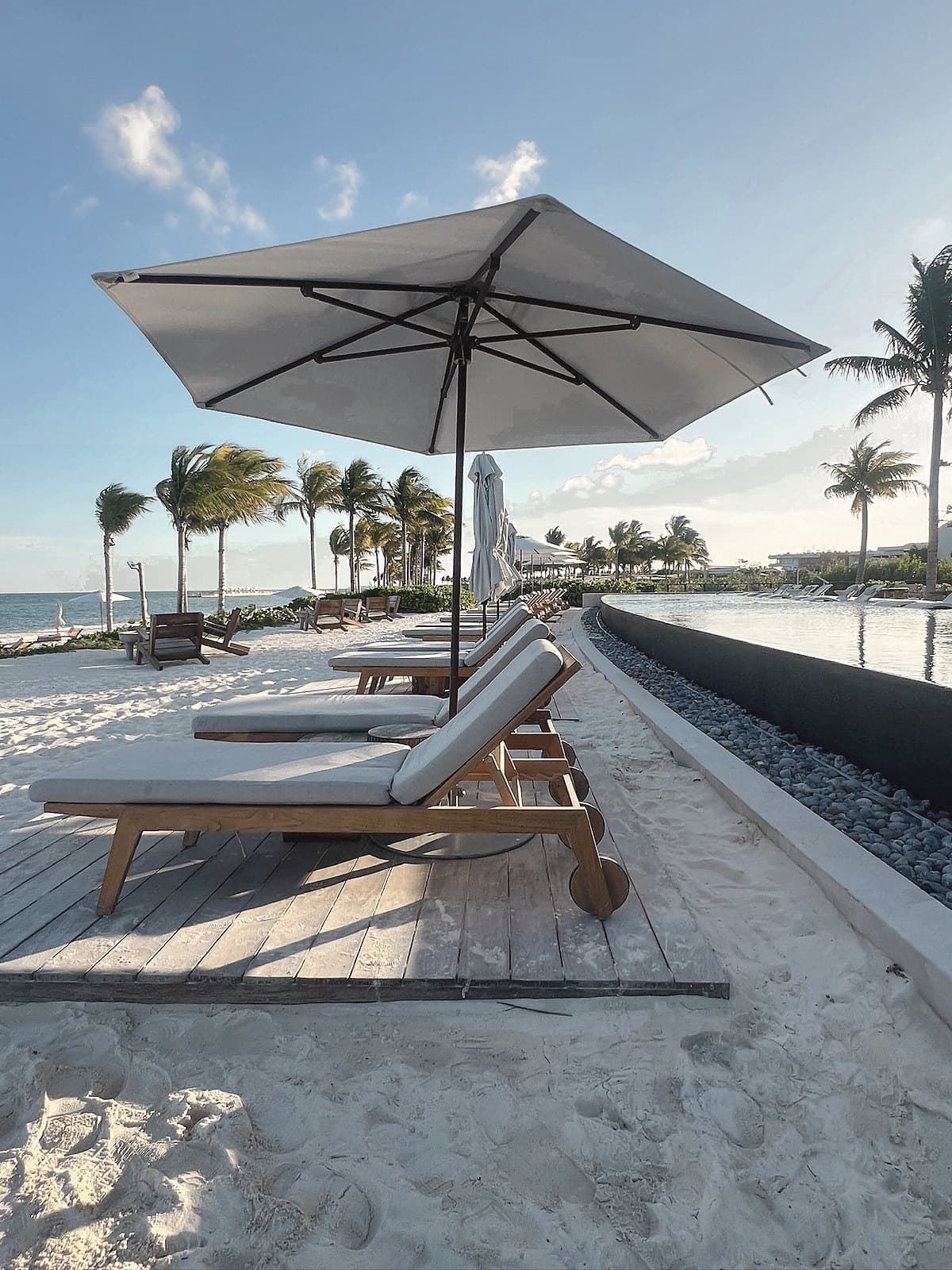 Teak loungers under a white umbrella on the sand beside the pool